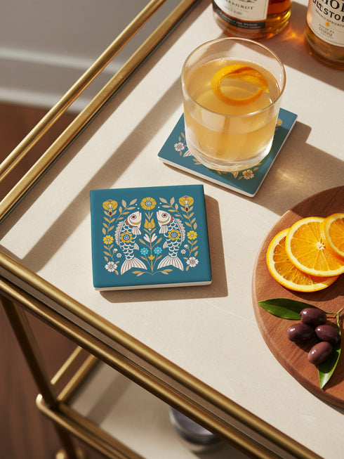Square ceramic coaster, 4x4 inches, with cork backing. Teal tile features a folk-art twin fish motif framed by florals in mustard, cream, and soft blue. Smooth printed surface; crisp, flat color artwork. Witchy zodiac-inspired Pisces coaster for mystical home décor, coffee table protection, and gifting. Square ceramic drink coaster with cork base, teal fish design.