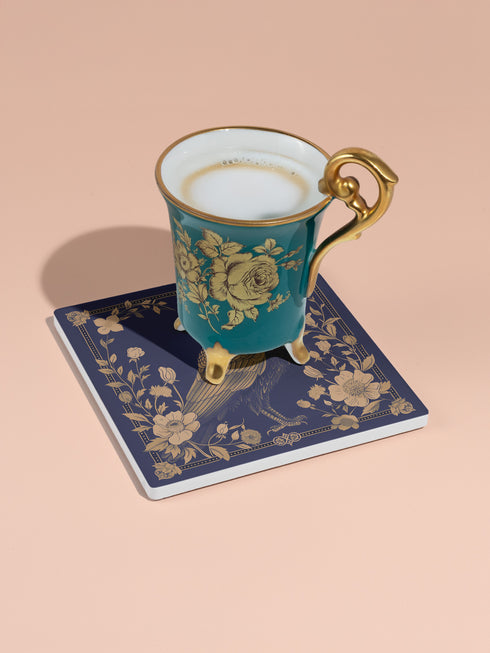 Square ceramic coaster featuring detailed illustration of a raven surrounded by golden florals and botanicals on navy background. Decorative border with dotted pattern frames the design.