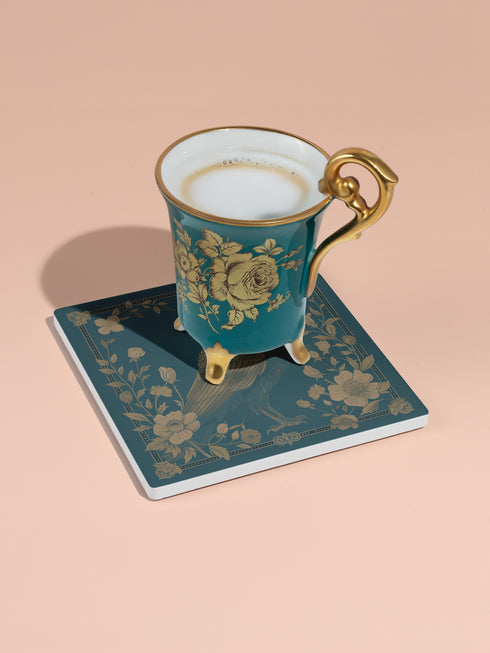 Square ceramic coaster featuring detailed gold raven illustration surrounded by botanical elements on emerald green background, with ornate border design and cork backing.