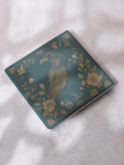 Square ceramic coaster featuring detailed gold raven illustration surrounded by botanical elements on emerald green background, with ornate border design and cork backing.