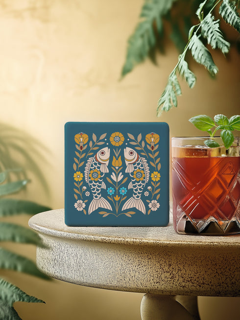 Square ceramic coaster, 4x4 inches, with cork backing. Teal tile features a folk-art twin fish motif framed by florals in mustard, cream, and soft blue. Smooth printed surface; crisp, flat color artwork. Witchy zodiac-inspired Pisces coaster for mystical home décor, coffee table protection, and gifting. Square ceramic drink coaster with cork base, teal fish design.