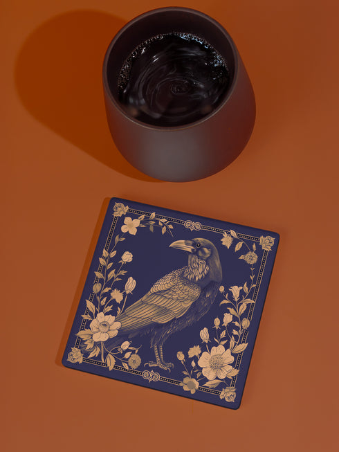 Square ceramic coaster featuring detailed illustration of a raven surrounded by golden florals and botanicals on navy background. Decorative border with dotted pattern frames the design.