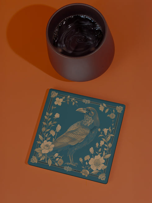 Square ceramic coaster featuring detailed gold raven illustration surrounded by botanical elements on emerald green background, with ornate border design and cork backing.
