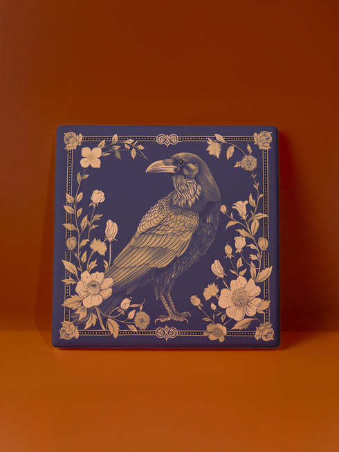 Square ceramic coaster featuring detailed illustration of a raven surrounded by golden florals and botanicals on navy background. Decorative border with dotted pattern frames the design.