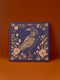 Square ceramic coaster featuring detailed illustration of a raven surrounded by golden florals and botanicals on navy background. Decorative border with dotted pattern frames the design.