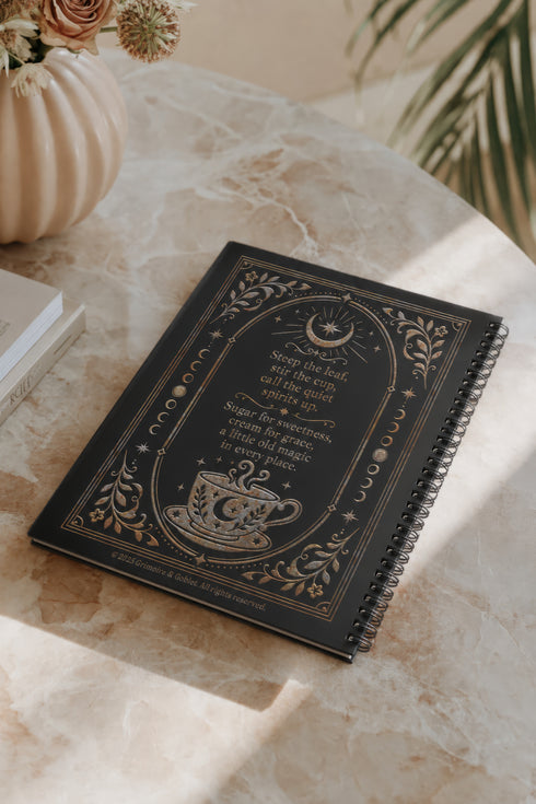 Spiral-bound recipe journal with a black cover and a metallic gold and silver design. The front reads Tea Leaves and Table Spells, Recipes for Sips, Sweets and Small Magic, framed by an arch, a crescent moon and star, moon phases, botanical sprigs, and a steaming teacup. The wire-O binding runs along one edge. Inside are blank fill-in recipe pages with lines for the recipe name, kitchen of origin, prep and cook time, servings, ingredients, and instructions.