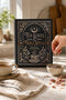 Spiral-bound recipe journal with a black cover and a metallic gold and silver design. The front reads Tea Leaves and Table Spells, Recipes for Sips, Sweets and Small Magic, framed by an arch, a crescent moon and star, moon phases, botanical sprigs, and a steaming teacup. The wire-O binding runs along one edge. Inside are blank fill-in recipe pages with lines for the recipe name, kitchen of origin, prep and cook time, servings, ingredients, and instructions.