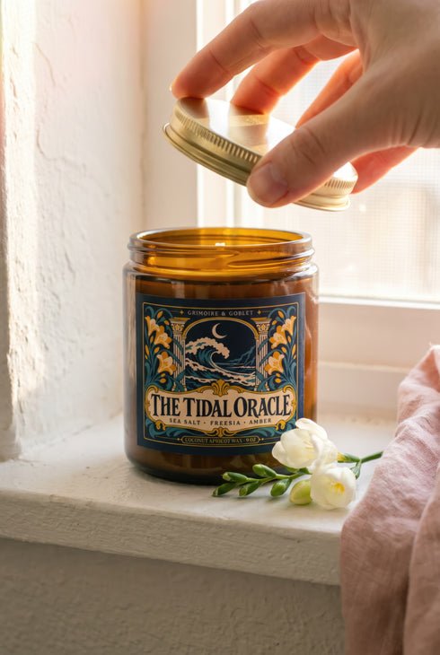 A 9 oz amber glass jar candle with a brushed gold screw lid and a wraparound deep navy label. The label shows a cresting wave beneath a crescent moon, framed by two classical columns and an arched window, with golden art nouveau freesias climbing both sides on teal stems, all inside a fine gold border. A cream banner reads The Tidal Oracle, with Grimoire & Goblet above and Sea Salt, Freesia, Amber and Coconut Apricot Wax, 9 oz below. A celestial coastal design.