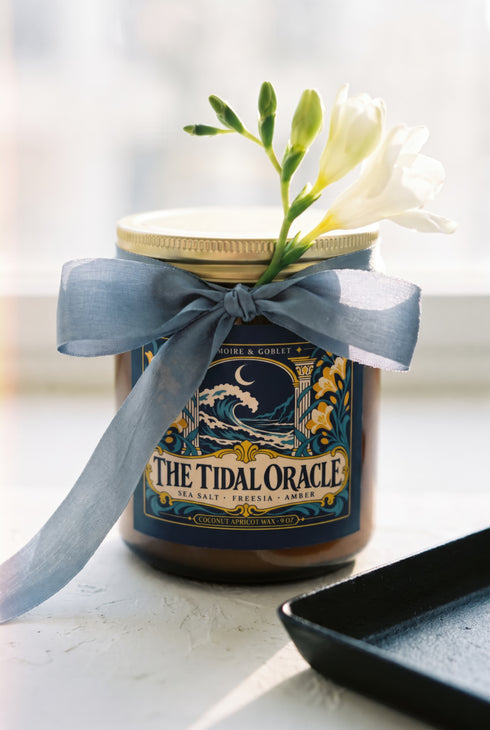 A 9 oz amber glass jar candle with a brushed gold screw lid and a wraparound deep navy label. The label shows a cresting wave beneath a crescent moon, framed by two classical columns and an arched window, with golden art nouveau freesias climbing both sides on teal stems, all inside a fine gold border. A cream banner reads The Tidal Oracle, with Grimoire & Goblet above and Sea Salt, Freesia, Amber and Coconut Apricot Wax, 9 oz below. A celestial coastal design.