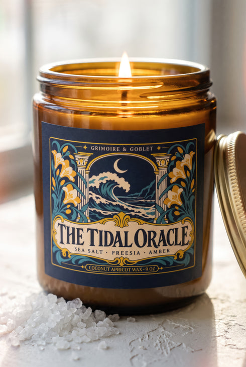 A 9 oz amber glass jar candle with a brushed gold screw lid and a wraparound deep navy label. The label shows a cresting wave beneath a crescent moon, framed by two classical columns and an arched window, with golden art nouveau freesias climbing both sides on teal stems, all inside a fine gold border. A cream banner reads The Tidal Oracle, with Grimoire & Goblet above and Sea Salt, Freesia, Amber and Coconut Apricot Wax, 9 oz below. A celestial coastal design.