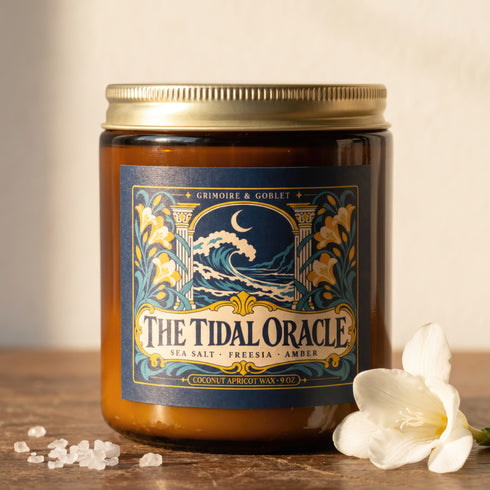 A 9 oz amber glass jar candle with a brushed gold screw lid and a wraparound deep navy label. The label shows a cresting wave beneath a crescent moon, framed by two classical columns and an arched window, with golden art nouveau freesias climbing both sides on teal stems, all inside a fine gold border. A cream banner reads The Tidal Oracle, with Grimoire & Goblet above and Sea Salt, Freesia, Amber and Coconut Apricot Wax, 9 oz below. A celestial coastal design.