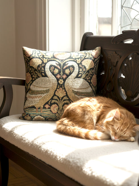 A square decorative throw pillow by Grimoire and Goblet featuring a woven jacquard tapestry design. The pattern displays two regal white swans facing each other with curved necks forming a heart shape, set against a dark background rich with intricate floral botany in beige, dusty rose, and sage green tones. The texture is thick and fabric-like, mimicking vintage needlepoint or Arts and Crafts style. The pillow is stuffed and sewn shut without a visible zipper.