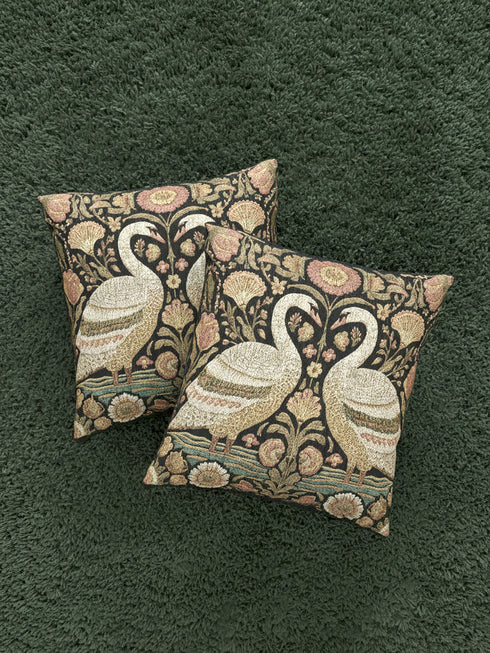 A square decorative throw pillow by Grimoire and Goblet featuring a woven jacquard tapestry design. The pattern displays two regal white swans facing each other with curved necks forming a heart shape, set against a dark background rich with intricate floral botany in beige, dusty rose, and sage green tones. The texture is thick and fabric-like, mimicking vintage needlepoint or Arts and Crafts style. The pillow is stuffed and sewn shut without a visible zipper.