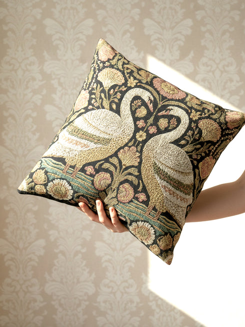 A square decorative throw pillow by Grimoire and Goblet featuring a woven jacquard tapestry design. The pattern displays two regal white swans facing each other with curved necks forming a heart shape, set against a dark background rich with intricate floral botany in beige, dusty rose, and sage green tones. The texture is thick and fabric-like, mimicking vintage needlepoint or Arts and Crafts style. The pillow is stuffed and sewn shut without a visible zipper.