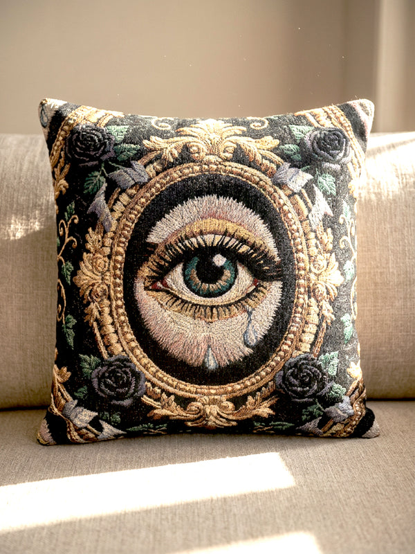 A square woven tapestry throw pillow by Grimoire and Goblet featuring a striking gothic design. The central motif is a large, detailed eye with a teal iris and light blue teardrops, set against pale skin. This eye is encased in an ornate, gold-toned baroque oval frame. The background is deep black, adorned with dark mauve roses and muted green foliage in the corners. The texture is a rich weave.