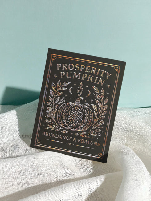 A 5x7 inch Prosperity Pumpkin ritual card shown on a flat surface. The card is made of thick, premium black card stock with a slight sheen. The front features an intricate, centrally-placed pumpkin illustration, ornately detailed with swirling patterns. The pumpkin and surrounding botanical elements, including leaves and cinnamon sticks, are rendered in a shimmering, gold and copper print. The words "Prosperity Pumpkin" and "Abundance & Fortune" are written in a serif font.