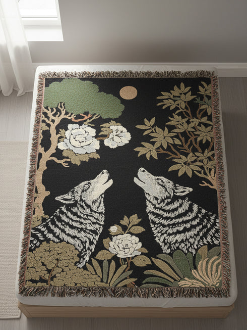 A high-contrast jacquard woven blanket featuring two wolves howling toward a central moon. The design is rendered in charcoal, sage green, and cream threads, showcasing intricate floral motifs like peonies and leafy trees. The textile has a visible woven texture with a multicolored fringe border. This Grimoire and Goblet piece displays a symmetrical, nature-inspired scene that functions as both a heavy throw and a wall tapestry, embodying a dark, botanical aesthetic.