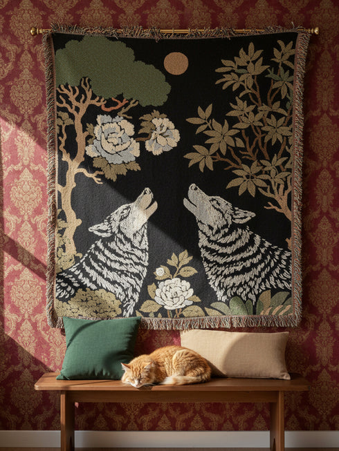A high-contrast jacquard woven blanket featuring two wolves howling toward a central moon. The design is rendered in charcoal, sage green, and cream threads, showcasing intricate floral motifs like peonies and leafy trees. The textile has a visible woven texture with a multicolored fringe border. This Grimoire and Goblet piece displays a symmetrical, nature-inspired scene that functions as both a heavy throw and a wall tapestry, embodying a dark, botanical aesthetic.