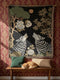 A high-contrast jacquard woven blanket featuring two wolves howling toward a central moon. The design is rendered in charcoal, sage green, and cream threads, showcasing intricate floral motifs like peonies and leafy trees. The textile has a visible woven texture with a multicolored fringe border. This Grimoire and Goblet piece displays a symmetrical, nature-inspired scene that functions as both a heavy throw and a wall tapestry, embodying a dark, botanical aesthetic.