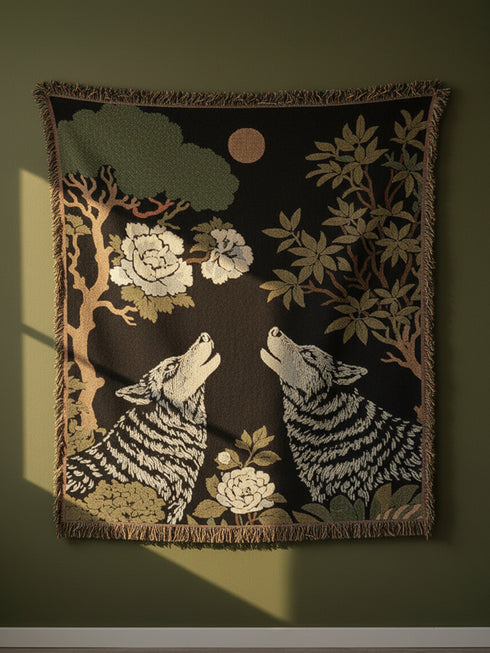A high-contrast jacquard woven blanket featuring two wolves howling toward a central moon. The design is rendered in charcoal, sage green, and cream threads, showcasing intricate floral motifs like peonies and leafy trees. The textile has a visible woven texture with a multicolored fringe border. This Grimoire and Goblet piece displays a symmetrical, nature-inspired scene that functions as both a heavy throw and a wall tapestry, embodying a dark, botanical aesthetic.