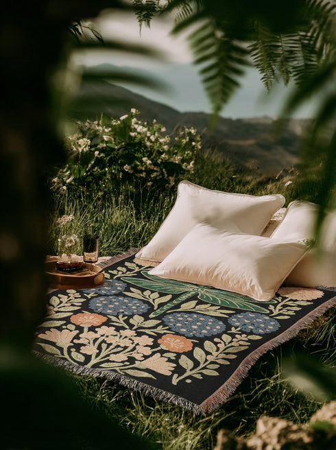 A Grimoire and Goblet Jacquard woven cotton blanket featuring a large central sage green dragonfly. The insect is surrounded by symmetrical clusters of slate blue hydrangeas and muted coral zinnia flowers against a black background. The blanket has a tactile woven texture and is finished with multicolor fringe edges. The design reflects a dark academia and vintage botanical aesthetic.