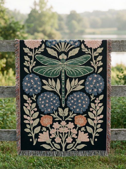 A Grimoire and Goblet Jacquard woven cotton blanket featuring a large central sage green dragonfly. The insect is surrounded by symmetrical clusters of slate blue hydrangeas and muted coral zinnia flowers against a black background. The blanket has a tactile woven texture and is finished with multicolor fringe edges. The design reflects a dark academia and vintage botanical aesthetic.