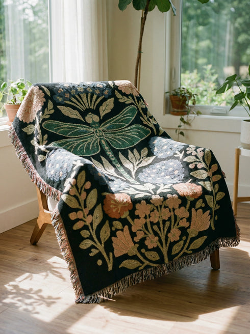 A Grimoire and Goblet Jacquard woven cotton blanket featuring a large central sage green dragonfly. The insect is surrounded by symmetrical clusters of slate blue hydrangeas and muted coral zinnia flowers against a black background. The blanket has a tactile woven texture and is finished with multicolor fringe edges. The design reflects a dark academia and vintage botanical aesthetic.