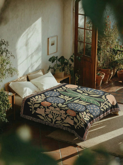 A Grimoire and Goblet Jacquard woven cotton blanket featuring a large central sage green dragonfly. The insect is surrounded by symmetrical clusters of slate blue hydrangeas and muted coral zinnia flowers against a black background. The blanket has a tactile woven texture and is finished with multicolor fringe edges. The design reflects a dark academia and vintage botanical aesthetic.
