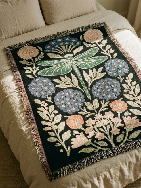 A Grimoire and Goblet Jacquard woven cotton blanket featuring a large central sage green dragonfly. The insect is surrounded by symmetrical clusters of slate blue hydrangeas and muted coral zinnia flowers against a black background. The blanket has a tactile woven texture and is finished with multicolor fringe edges. The design reflects a dark academia and vintage botanical aesthetic.
