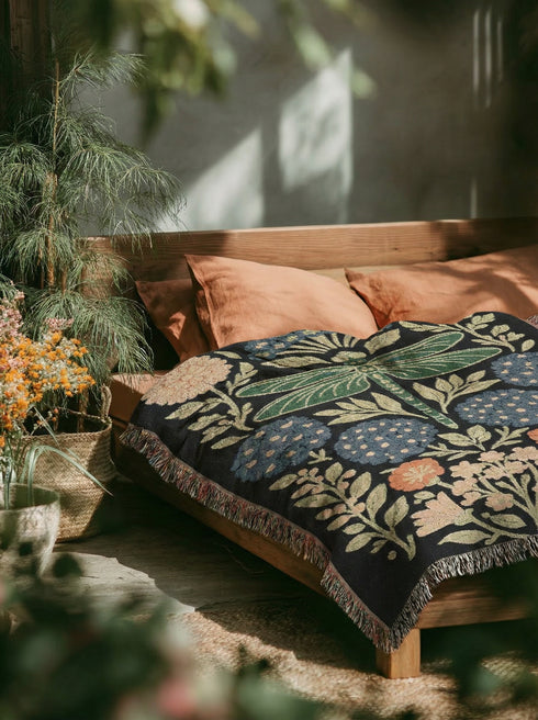 A Grimoire and Goblet Jacquard woven cotton blanket featuring a large central sage green dragonfly. The insect is surrounded by symmetrical clusters of slate blue hydrangeas and muted coral zinnia flowers against a black background. The blanket has a tactile woven texture and is finished with multicolor fringe edges. The design reflects a dark academia and vintage botanical aesthetic.