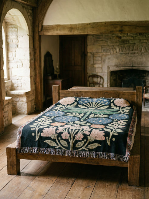 A Grimoire and Goblet Jacquard woven cotton blanket featuring a large central sage green dragonfly. The insect is surrounded by symmetrical clusters of slate blue hydrangeas and muted coral zinnia flowers against a black background. The blanket has a tactile woven texture and is finished with multicolor fringe edges. The design reflects a dark academia and vintage botanical aesthetic.