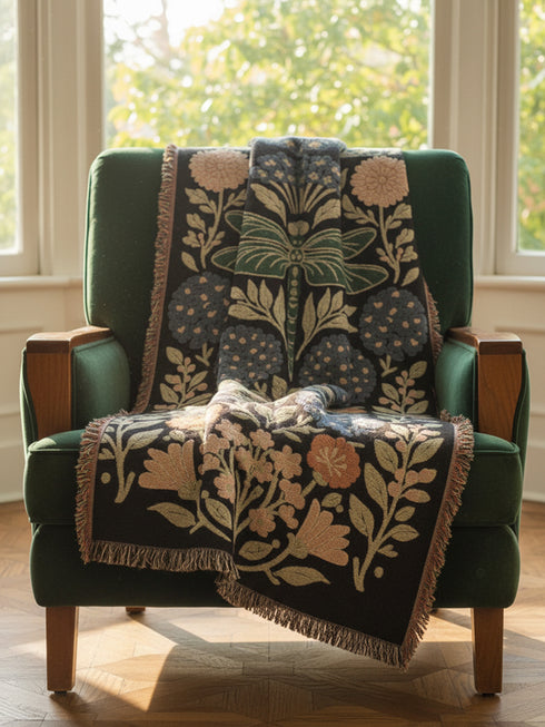 A Grimoire and Goblet Jacquard woven cotton blanket featuring a large central sage green dragonfly. The insect is surrounded by symmetrical clusters of slate blue hydrangeas and muted coral zinnia flowers against a black background. The blanket has a tactile woven texture and is finished with multicolor fringe edges. The design reflects a dark academia and vintage botanical aesthetic.