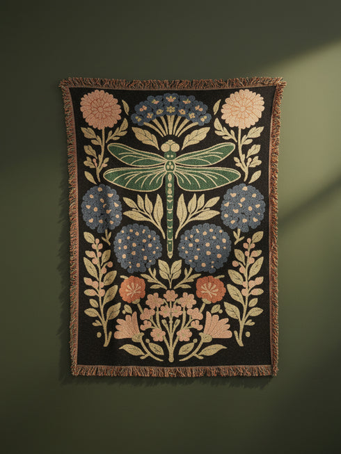 A Grimoire and Goblet Jacquard woven cotton blanket featuring a large central sage green dragonfly. The insect is surrounded by symmetrical clusters of slate blue hydrangeas and muted coral zinnia flowers against a black background. The blanket has a tactile woven texture and is finished with multicolor fringe edges. The design reflects a dark academia and vintage botanical aesthetic.