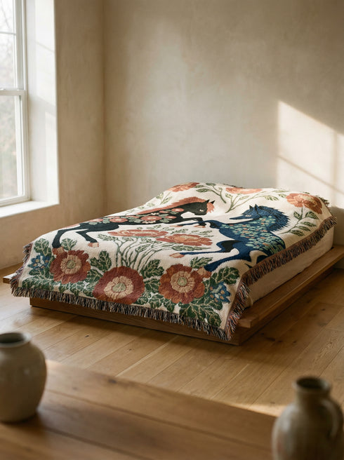 A woven cotton throw blanket by Grimoire and Goblet featuring two rearing horses facing each other in a symmetrical folk art design. One horse is charcoal black with rust floral detailing; the other is navy blue with teal floral detailing. They are framed by large rust-colored peonies and green leafy vines on a cream background. The blanket features a multicolor fringe edge. Perfect for dark academia and cottagecore aesthetics.