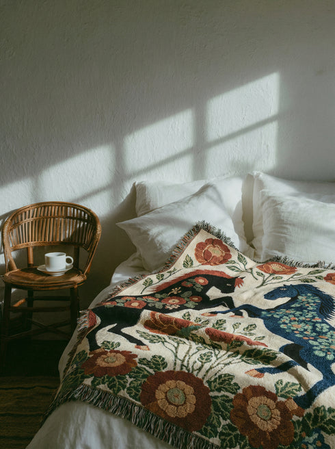A woven cotton throw blanket by Grimoire and Goblet featuring two rearing horses facing each other in a symmetrical folk art design. One horse is charcoal black with rust floral detailing; the other is navy blue with teal floral detailing. They are framed by large rust-colored peonies and green leafy vines on a cream background. The blanket features a multicolor fringe edge. Perfect for dark academia and cottagecore aesthetics.