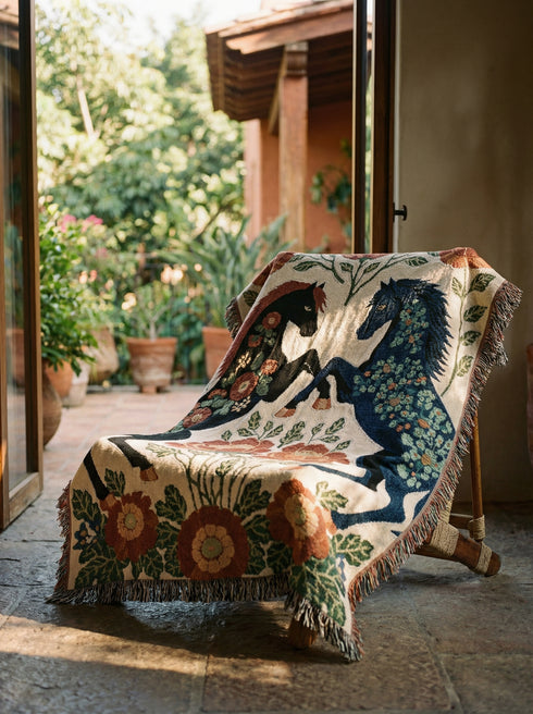 A woven cotton throw blanket by Grimoire and Goblet featuring two rearing horses facing each other in a symmetrical folk art design. One horse is charcoal black with rust floral detailing; the other is navy blue with teal floral detailing. They are framed by large rust-colored peonies and green leafy vines on a cream background. The blanket features a multicolor fringe edge. Perfect for dark academia and cottagecore aesthetics.