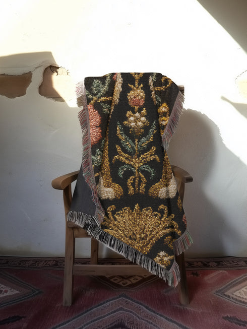 A rectangular, 100% woven cotton throw blanket with a detailed jacquard design. The throw features a symmetrical, vertical pattern of two howling wolves in profile, rendered in shades of beige, gold, and brown against a solid black background. The wolves are surrounded by intricate floral and foliage motifs. The thick, textured weave is visible. The blanket is finished on all four sides with a two-inch, multi-colored fringe that includes black, white, red, and gold threads.