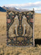A rectangular, 100% woven cotton throw blanket with a detailed jacquard design. The throw features a symmetrical, vertical pattern of two howling wolves in profile, rendered in shades of beige, gold, and brown against a solid black background. The wolves are surrounded by intricate floral and foliage motifs. The thick, textured weave is visible. The blanket is finished on all four sides with a two-inch, multi-colored fringe that includes black, white, red, and gold threads.