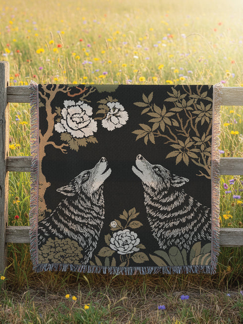 A high-contrast jacquard woven blanket featuring two wolves howling toward a central moon. The design is rendered in charcoal, sage green, and cream threads, showcasing intricate floral motifs like peonies and leafy trees. The textile has a visible woven texture with a multicolored fringe border. This Grimoire and Goblet piece displays a symmetrical, nature-inspired scene that functions as both a heavy throw and a wall tapestry, embodying a dark, botanical aesthetic.