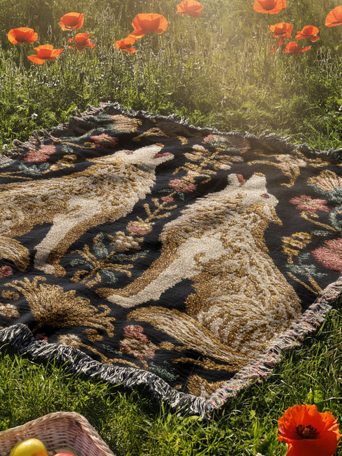 A rectangular, 100% woven cotton throw blanket with a detailed jacquard design. The throw features a symmetrical, vertical pattern of two howling wolves in profile, rendered in shades of beige, gold, and brown against a solid black background. The wolves are surrounded by intricate floral and foliage motifs. The thick, textured weave is visible. The blanket is finished on all four sides with a two-inch, multi-colored fringe that includes black, white, red, and gold threads.