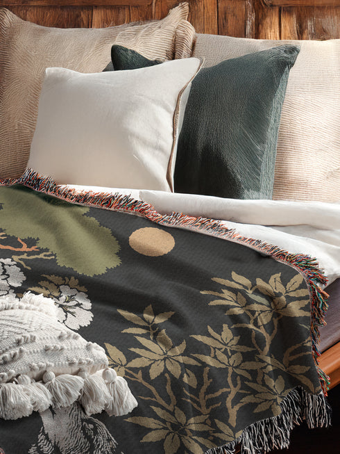 A high-contrast jacquard woven blanket featuring two wolves howling toward a central moon. The design is rendered in charcoal, sage green, and cream threads, showcasing intricate floral motifs like peonies and leafy trees. The textile has a visible woven texture with a multicolored fringe border. This Grimoire and Goblet piece displays a symmetrical, nature-inspired scene that functions as both a heavy throw and a wall tapestry, embodying a dark, botanical aesthetic.
