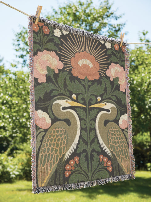 Jacquard woven cotton throw blanket, woven rather than printed, shown flat. Two mirrored great blue herons flank a central radial sunburst, surrounded by botanical blooms and scrollwork. The charcoal black ground carries antique gold ochre sunbeams, blush pink blossoms, deep forest green leaves, muted rust berry clusters, and cream toned herons. A lush multi-colored fringe of black, ochre, and sage threads edges the blanket, and the reverse shows an inverted charcoal grey version of the same design.