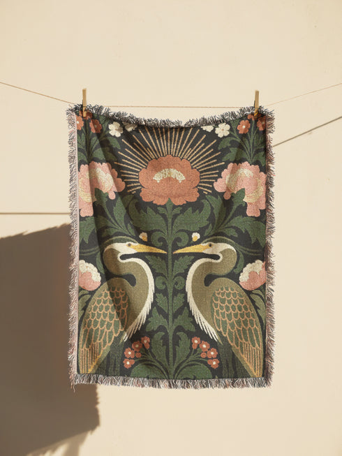Jacquard woven cotton throw blanket, woven rather than printed, shown flat. Two mirrored great blue herons flank a central radial sunburst, surrounded by botanical blooms and scrollwork. The charcoal black ground carries antique gold ochre sunbeams, blush pink blossoms, deep forest green leaves, muted rust berry clusters, and cream toned herons. A lush multi-colored fringe of black, ochre, and sage threads edges the blanket, and the reverse shows an inverted charcoal grey version of the same design.