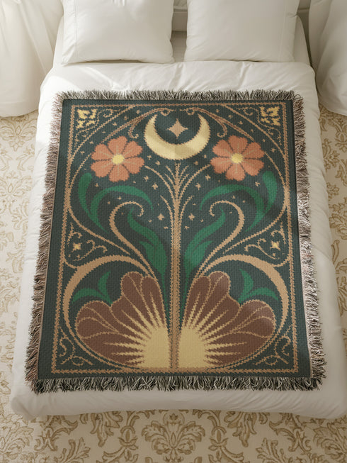 A luxurious, intricately woven jacquard blanket from Grimoire and Goblet. The design is a symmetrical Art Nouveau botanical pattern on a dark teal background. A central stem with green leaves swirls upwards, flanked by two large, rust-orange flowers. At the top, a golden crescent moon and star sparkle. A fan-like, sun-inspired motif rests at the base. The entire piece, crafted from 100% cotton, is framed by a decorative gold border and a multi-colored fringe.
