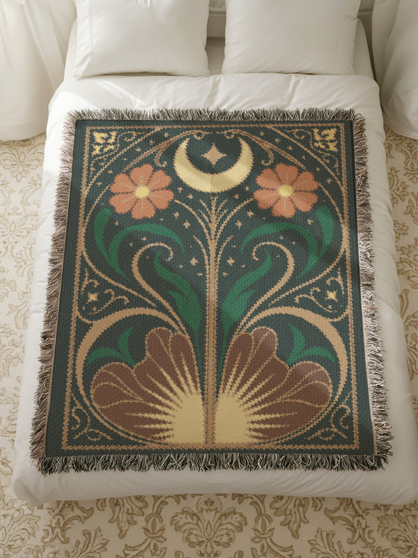 A luxurious, intricately woven jacquard blanket from Grimoire and Goblet. The design is a symmetrical Art Nouveau botanical pattern on a dark teal background. A central stem with green leaves swirls upwards, flanked by two large, rust-orange flowers. At the top, a golden crescent moon and star sparkle. A fan-like, sun-inspired motif rests at the base. The entire piece, crafted from 100% cotton, is framed by a decorative gold border and a multi-colored fringe.
