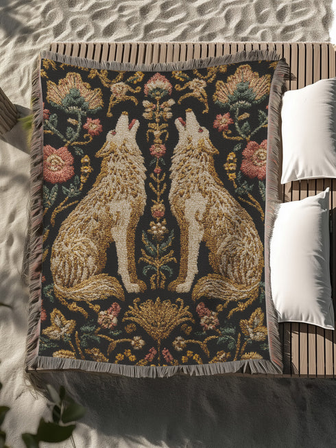 A rectangular, 100% woven cotton throw blanket with a detailed jacquard design. The throw features a symmetrical, vertical pattern of two howling wolves in profile, rendered in shades of beige, gold, and brown against a solid black background. The wolves are surrounded by intricate floral and foliage motifs. The thick, textured weave is visible. The blanket is finished on all four sides with a two-inch, multi-colored fringe that includes black, white, red, and gold threads.