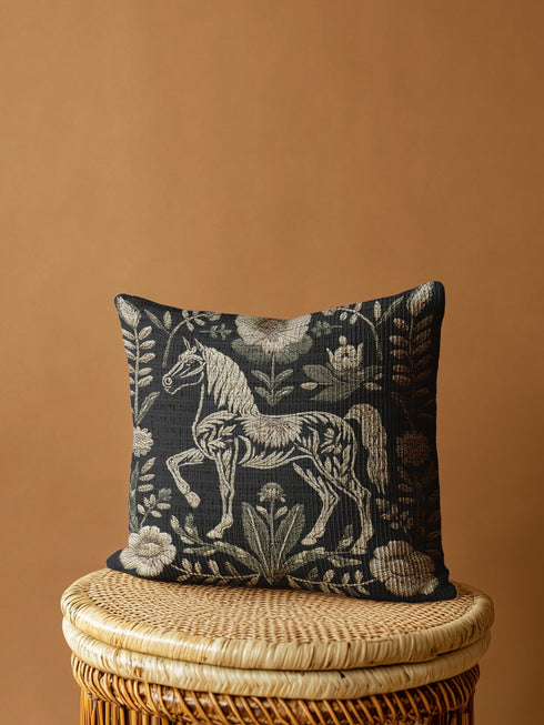 A premium, artisan-woven accent pillow from Grimoire and Goblet. The pillow is 17x18 and features a highly detailed, double-sided jacquard design on a deep black background. The artwork, woven in neutral gold, ecru, and muted olive threads, depicts a mystical, folkloric horse. This central horse figure is surrounded by a lush, symmetrical border of botanical flowers and leaves. The style is evocative of a medieval tapestry, perfect for dark academia, gothic, or arcane-themed decor.