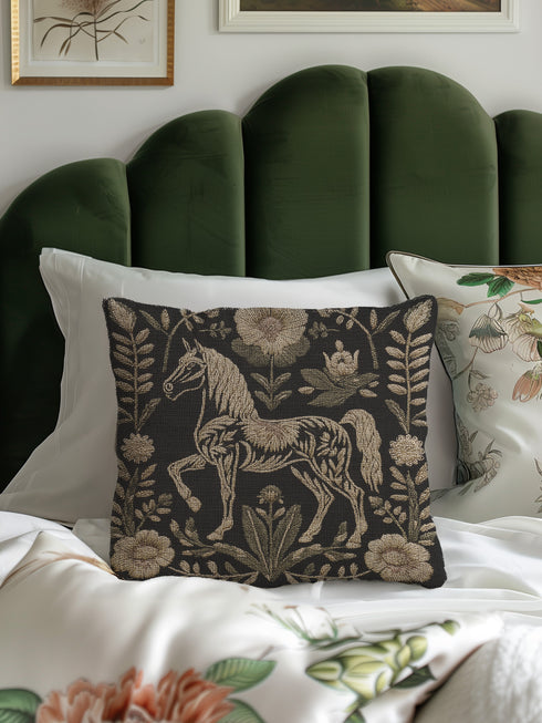 A premium, artisan-woven accent pillow from Grimoire and Goblet. The pillow is 17x18 and features a highly detailed, double-sided jacquard design on a deep black background. The artwork, woven in neutral gold, ecru, and muted olive threads, depicts a mystical, folkloric horse. This central horse figure is surrounded by a lush, symmetrical border of botanical flowers and leaves. The style is evocative of a medieval tapestry, perfect for dark academia, gothic, or arcane-themed decor.