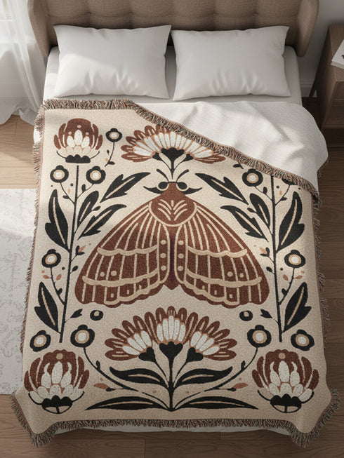 Grimoire and Goblet woven jacquard cotton blanket / wall tapestry featuring a centered folk-art moth framed by symmetrical wildflowers and leafy stems. Natural oatmeal-beige base with rust/terracotta and cocoa-brown moth detail, charcoal-black botanicals, and warm ivory highlights. Finished with a 2-inch multicolor fringe and a reversible color-negative back. Available in 52x37, 50x60, and 60x80 inches as a throw, bed cover, or statement wall hanging.