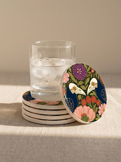 A square drink coaster with a high-gloss finish and natural cork backing. The centralized, symmetrical design features two detailed, cream-colored avian bird skulls facing inward. They are framed by lush botanical flora, including a large purple flower, blush pink blooms, rust-red roses, and green ferns, all set against a deep navy blue background with gold linework accents. Functional gothic, whimsigoth, and dark academia drinkware designed by the Grimoire and Goblet brand.